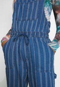 Salopette bleu denim à rayures blanches verticales portée sur un t-shirt tie-dye multicolore à manches longues, mains dans les poches.