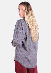 Femme avec de longs cheveux blonds ondulés portant une chemise bleue ample à motifs et un pantalon rouge, jetant un coup d'œil par-dessus son épaule devant un fond uni.