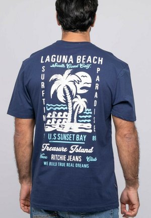 Homme portant un t-shirt bleu marine avec des motifs et textes de surf en blanc et bleu clair faisant référence à Laguna Beach, des palmiers, des vagues et « Treasure Island ».
