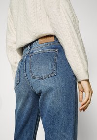 Jeans de ganga azul de cintura alta com uma textura ligeiramente desgastada, apresentando dois bolsos traseiros e um emblema de pele castanha na cintura.