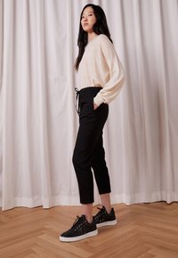 Béžový dlouhý rukáv, černé kalhoty na šňůrku a černé tenisky se zlatými akcenty. Texturované materiály, uvolněný střih, viditelné boční kapsy.
