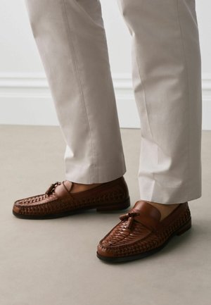 Mand iført lyse beige bukser og brune flettede læderloafer med kvaster, stående på en glat beige gulv nær en hvid vægpanel.