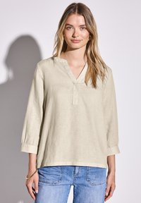 Camisetta a maniche lunghe color beige chiaro con una texture simile al lino, dotata di scollatura a V e vestibilità ampia, abbinata a jeans blu.