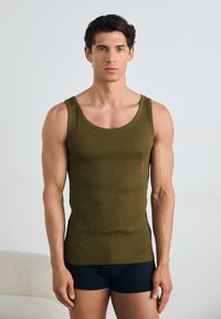 Olivgrünes, geripptes Tanktop mit breiten Trägern, schlanker Passform und kleinem roten Logo auf der Vorderseite. Model trägt schwarze Shorts.