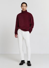 Maglione a collo alto bordeaux con polsi a coste, abbinato a pantaloni bianchi e stivali neri alla caviglia, in piedi di fronte a uno sfondo neutro.