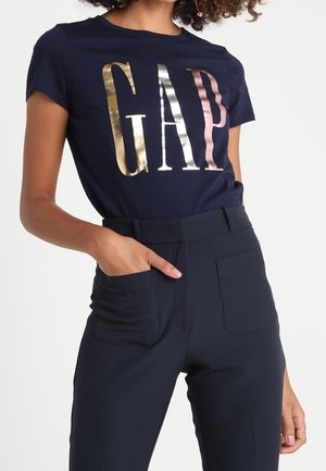 Kobieta w granatowym t-shircie z błyszczącymi złotymi, srebrnymi i różowymi literami „GAP”, w połączeniu z wysokim stanem ciemnogranatowych spodni z przednimi kieszeniami.