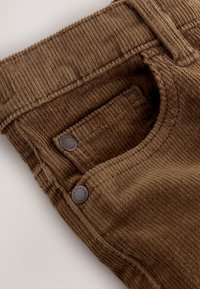 Pantalon en velours côtelé marron avec nervures verticales, doté de deux boutons en métal et d'une poche latérale. Texture douce et ceinture élastique.