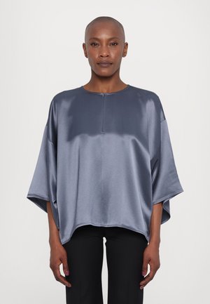 Graues Satin-Übergröße-Oberteil mit kurzen Ärmeln, runder Ausschnitt und einer kleinen Knopfleiste vorne, kombiniert mit schwarzen Hosen. Glatte Textur und lockerer Schnitt.