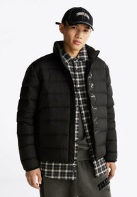 Czarna kurtka puchowa z pikowaną teksturą i detalem logo, noszona na szarej bluzie i czarno-białej koszuli w kratę. Czapka z logo.