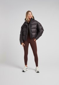 Donna bionda che indossa un giubbotto di piumino nero, leggings marroni e sneakers bianche, in piedi con una mano in tasca contro uno sfondo grigio neutro.