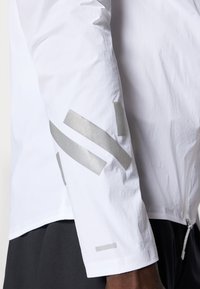 Giacca leggera bianca in tessuto testurizzato, con dettagli riflettenti argentati sulla manica sinistra e chiusura con zip.