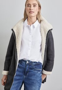 Giacca nera con colletto e foderato in ecopelliccia color crema, frontale con zip, indossata sopra una camicia bianca e jeans blu.