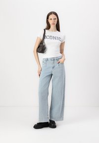 Balta marškinėliai su mėlynu užrašu "ICONIC", derinami su šviesiai mėlynomis plačiomis džinsomis ir juodais stambiais batais. Modelis laiko tamsią rankinę.