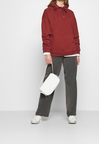 Felpa rossa con tasca frontale e cappuccio regolabile, abbinata a pantaloni larghi grigi e una borsa intrecciata bianca. Sneakers bianche completano il look.
