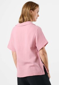 Rosa kurzärmliges Shirt aus strukturiertem Stoff mit weichem Kragen und seitlichen Schlitzen, von hinten an einem Modell präsentiert.