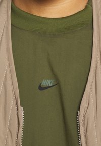 Zelené tričko Nike s malým logem na hrudi pod otevřeným béžovým kabátem s viditelným prošitím a zipem.