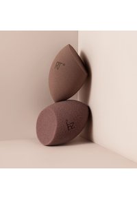 Deux éponges à maquillage marron avec le logo "RT", placées debout et couchées contre un mur beige avec un angle et une surface.