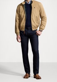 Khaki bomberjack, marineblauwe kabelgebreide trui, donkere spijkerbroek, bruine loafers. Opvallend logo op de borst; gladde stof en een getailleerde pasvorm.