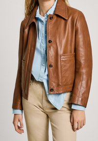 Giacca di pelle marrone con colletto, chiusura a bottoni e due tasche anteriori. Indossata sopra una camicia azzurro chiaro con bottoni. Tessuto liscio.