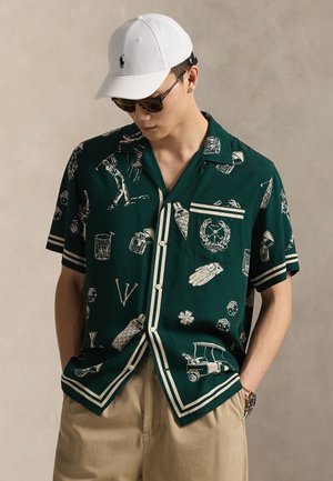 Mladý muž oblečený v tmavozelenej krátkosleevej košeli s potlačou golfovej témy, béžové nohavice, biela čiapka, slnečné okuliare a náramkové hodinky, ruky v vreckách.