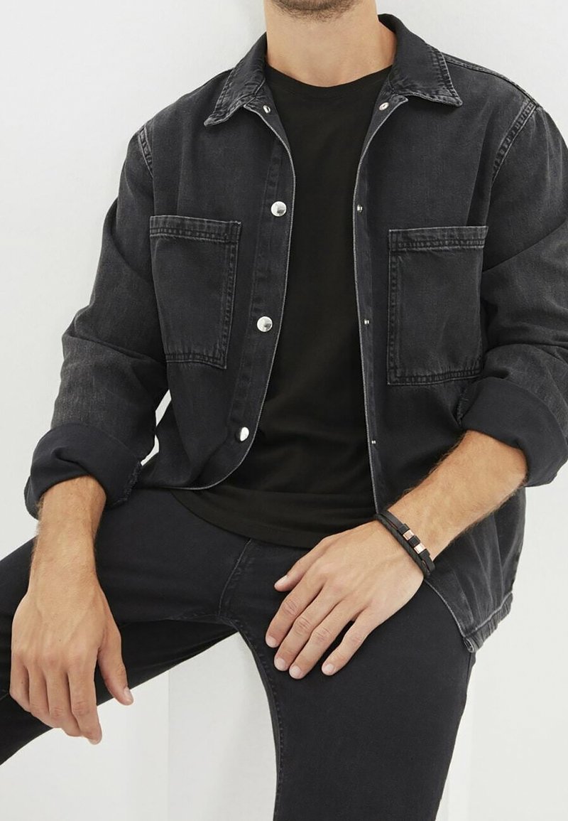 Chaqueta de denim negra con botones plateados, dos bolsillos y puños enrollados, llevada sobre una camiseta negra y combinada con jeans negros.
