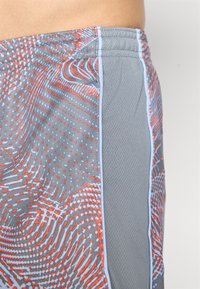 Nærbilde av grå treningsshorts med oransje og lyseblå abstrakte prikker og virvelmønstre, som viser teksturert stoff og elastisk livlinning.