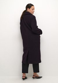 Lungo cappotto viola con una finitura testurizzata, dotato di colletto, spacchi laterali e maniche ampie, abbinato a pantaloni neri e sandali con tacco.