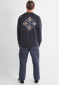 T-shirt noir à manches longues pour homme avec un graphisme coloré au dos, associé à un pantalon cargo bleu marine ample. Des baskets noires complètent le look.