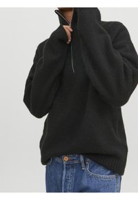 Osoba nosząca luźny czarny sweter z dzianiny z półzamkiem przy kołnierzu i niebieskie dżinsy, poprawiająca zamek przy szyi na jednolitym tle.