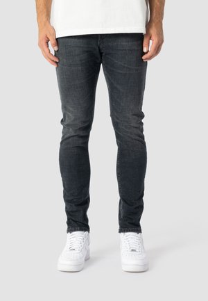 Mann trägt eng anliegende dunkelgraue Jeans und weiße Sneakers, steht vor einem einfarbigen hellgrauen Hintergrund.