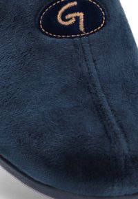 Zapato de ante azul marino con textura suave, detalles de costura y un logo bordado en un parche circular. Diseño de punta redondeada.