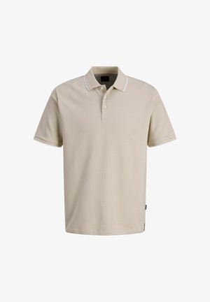 Ragazzo biondo che sorride, in piedi con le mani rilassate, indossa una polo beige a maniche corte, pantaloni scuri e sneakers bianche su sfondo bianco semplice.