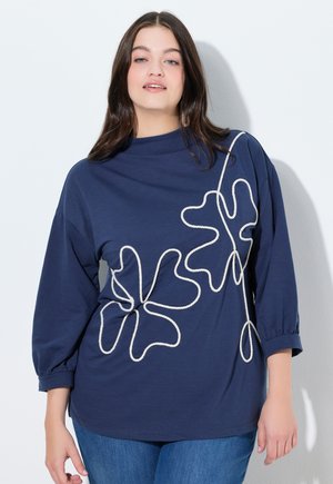 Giovane donna con capelli castani lunghi che indossa una maglia blu navy con motivi floreali astratti bianchi a linee e jeans blu, in piedi contro un muro bianco.