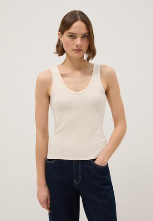 Femme aux cheveux bruns courts portant un haut beige côtelé sans manches et un jean bleu foncé, debout avec une main dans la poche sur un fond uni.