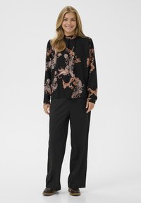 Mosolygó nő hosszú szőke hajjal, fekete paisley mintás blúzban, laza fekete nadrágban és sötétbarna cipőben, fehér háttér előtt.