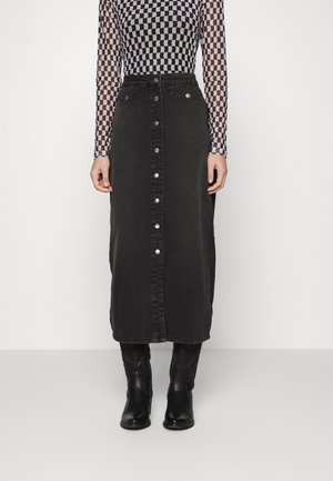 Femme portant une jupe midi en denim noire boutonnée devant, des bottes de cow-boy noires et un haut à manches longues à carreaux noirs et blancs.
