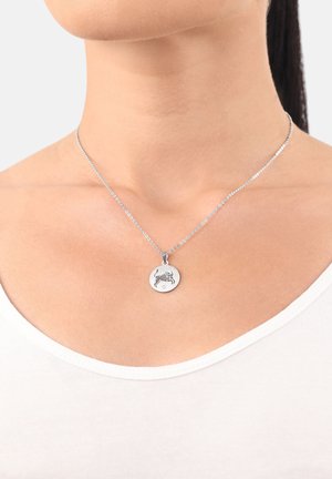 Collana d'argento con pendente tondo che presenta un simbolo di toro rialzato, indossata attorno al collo di una donna, su un top bianco e sfondo chiaro.