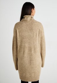 Beige kötött pulóver magas gallérral, túlméretezett fazonnal és bordás ujjrész részletekkel. Puha textúrájú, a comb közepéig ér.