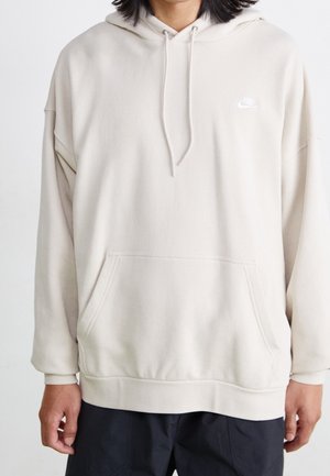 Person trägt einen hellbeigen Nike-Hoodie mit Kordelzug und Fronttasche, kombiniert mit dunklen Hosen, vor einem einfarbigen Hintergrund.