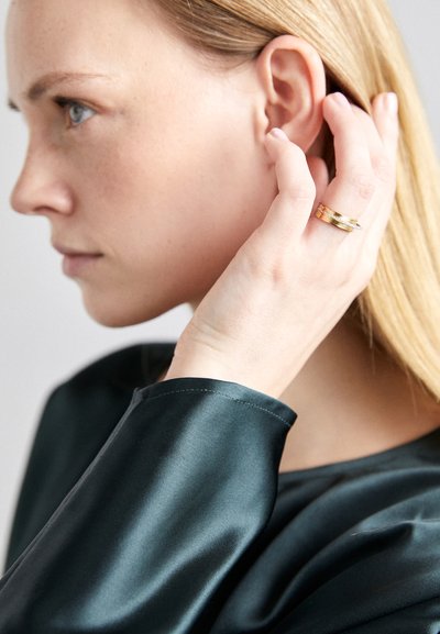 Anillo de oro con tres bandas entrelazadas, llevado en la mano derecha. El fondo presenta una superficie lisa de color claro y la persona lleva una blusa oscura de satén.