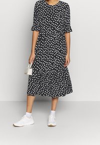 Mustat kukkakuosiset mekko lyhyillä hihoilla, röyhelöhelma ja valkoisia kukkia. Yhdistetty valkoisiin lenkkareihin ja pieneen valkoiseen laukkuihin.