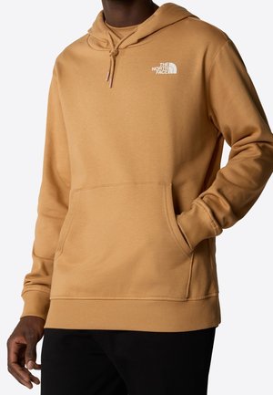 Mann trägt eine beige The North Face Kapuzenjacke mit Fronttasche, Kordelzug an der Kapuze und Logo auf der linken Brustseite, kombiniert mit schwarzen Hosen.