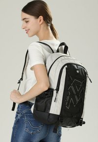 Zaino grigio e nero con spallacci imbottiti, design con cordino elastico frontale e tasca laterale in rete. Presenta una chiusura con cerniera e un tessuto resistente.