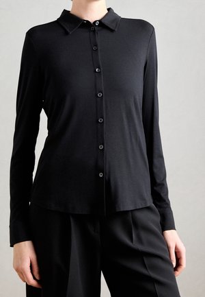 Personne portant une chemise noire ajustée à manches longues avec col et boutons, accompagnée d'un pantalon noir ample, sur un fond clair uni.