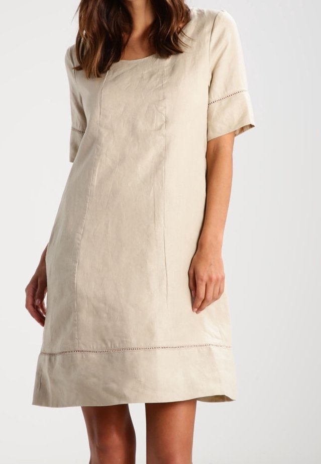 Femme portant une robe beige à manches courtes, longueur genou, avec des détails de coutures subtiles et un ourlet décoratif.