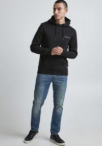Felpa nera con cappuccio, dotata di tasca frontale e stringhe, abbinata a jeans blu sbiaditi e sneakers nere. Design semplice, senza motivi visibili.