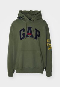 Hoodie vert avec logo "GAP" en navy et rouge, poche avant, capuche avec cordon de serrage, et texte brodé jaune sur la manche gauche. Texture de tissu douce.