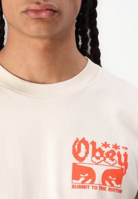 Lys beige bomull t-skjorte med et dristig rødt grafisk trykk med teksten "Obey" og "Submit to the Rhythm" på venstre brystområde.