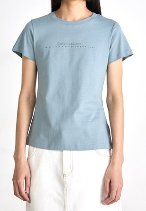 Person wearing a light blue Calvin Klein Jeans t-shirt and a white skirt, standing against a plain white background.