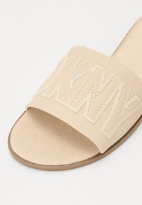 Beige kanvasu slaidu sandales ar platām siksnām, kas dekorētas ar izceltām baltām izšūtām burtiem. Gluda zole ar noapaļotu formu.
